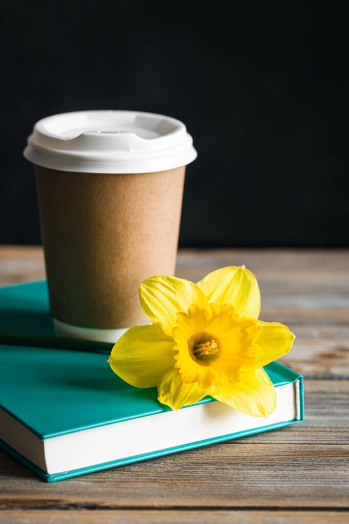 Paper cup, book and yellow flower on a blurred wooden background.
